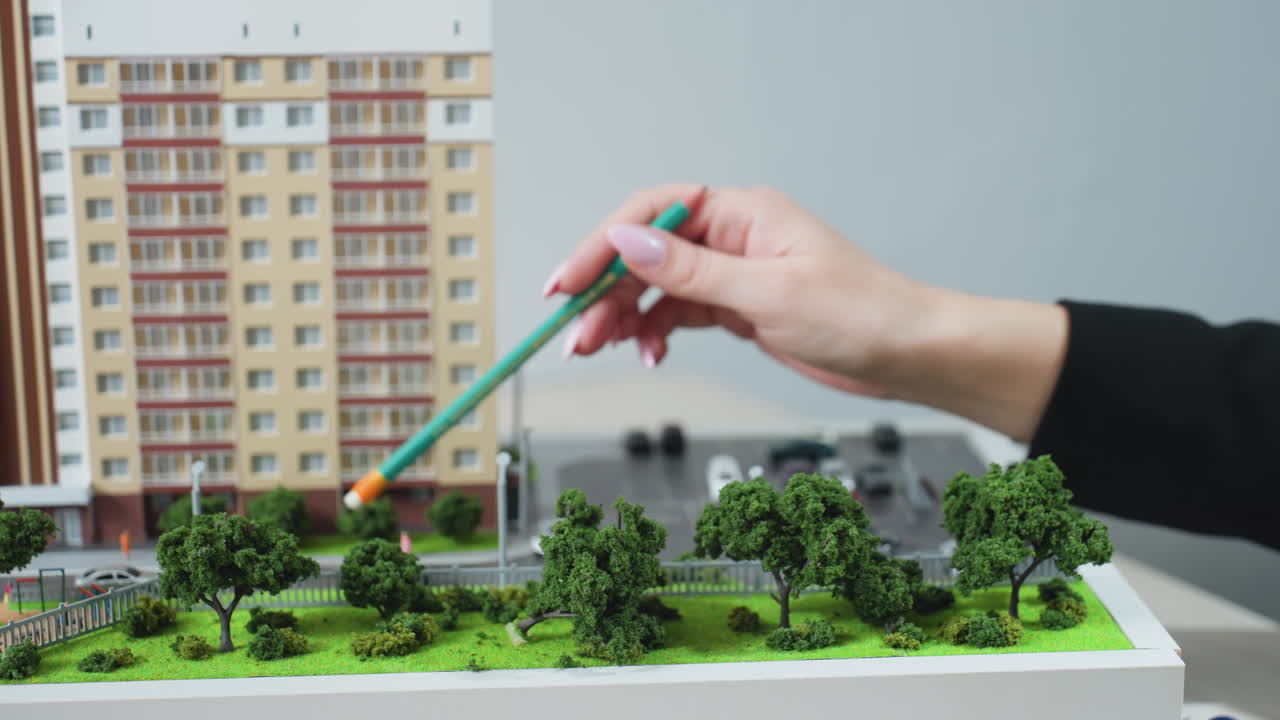 Close up of realtor hand holding pencil and pointing at miniature building model with visible trees and parked cars, suggesting property planning with car key nearby on desk