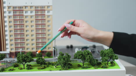 close up of realtor hand holding pencil and pointing at miniature building model with visible trees and parked cars, suggesting property planning with car key nearby on desk