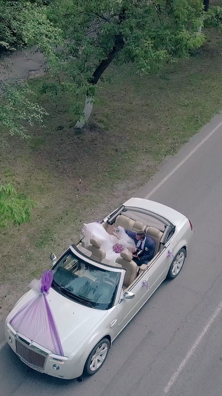 camera rises over bride in white dress with purple bouquet lying on husband knees in wedding convertible