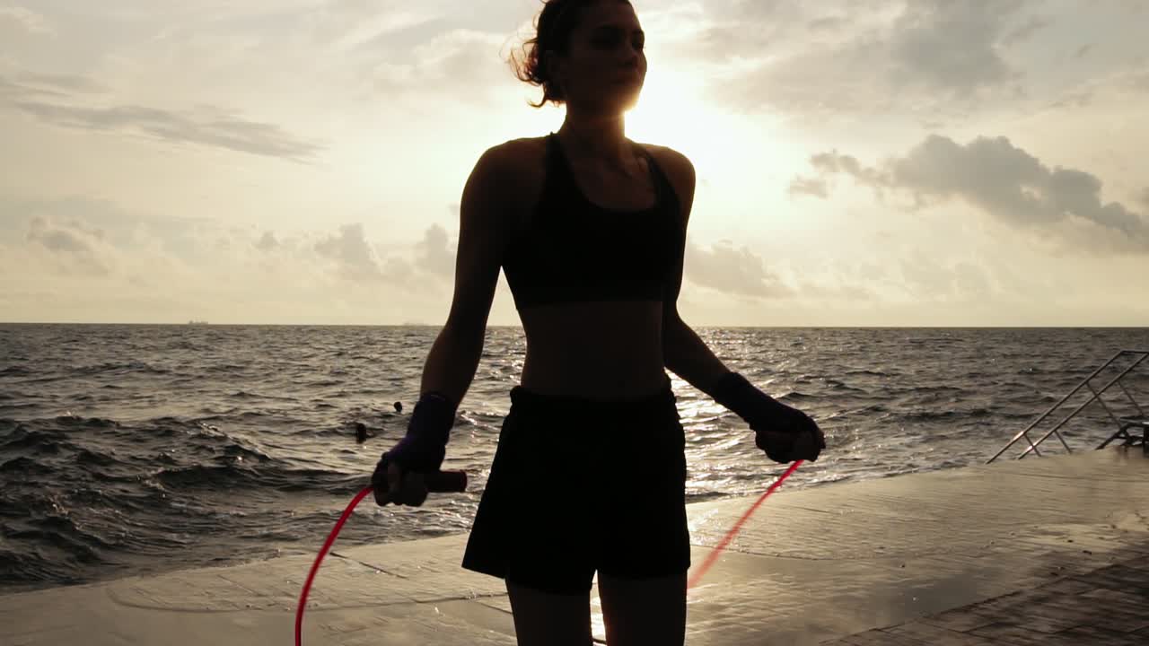 Closeup view of a young woman working out on the jump rope against the son by the beach in Slow Motion. Girl jumping on a skipping
