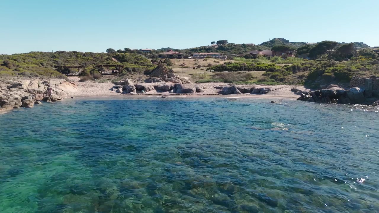 Una vista aerea si avvicina a una spiaggia rocciosa sarda con mare azzurro cristallino e colline costiere.