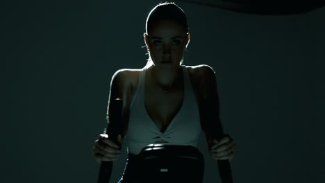 frontal waist-up shot of a woman gripping the handlebar while cycling, as the camera glides slightly from left to right. cinematic backlight reveals sweat on her neckline and sculpted upper body