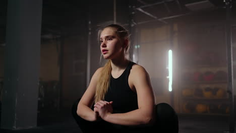 a young slender woman prepares and warms up before training. hitching and stretching muscles after a tedious hard workout in the dark interior of the fitness room