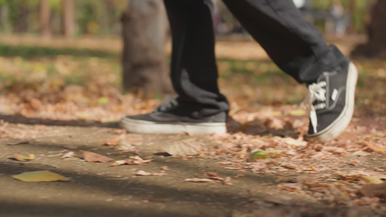 Chaussures d'adulte marchant à travers les feuilles mortes sur un chemin de parc, pas régulier en baskets noires, lumière dorée de l'après-midi, jeans décontracté et allure détendue, ambiance de promenade familiale tranquille et craquement vif des feuilles.
