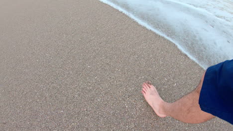 Man's-feet-walking-in-the-sand-on-a-cloudy-day,-along-edge-of-surf