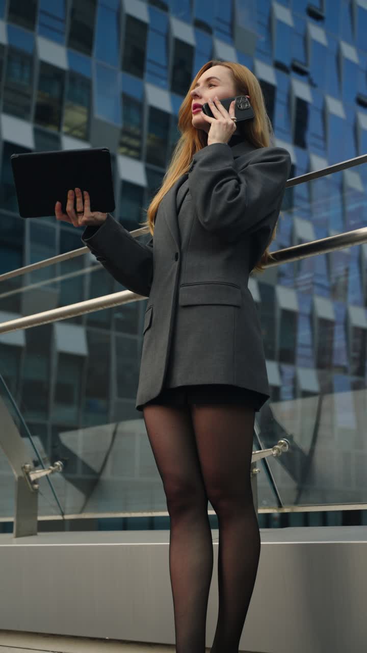 Professional businesswoman stands on a terrace, holding a tablet and talking seriously on her phone, surrounded by modern glass office buildings - parallax shot