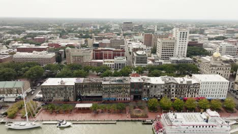 Drone-of-Savannah-Georgia-riverfront-area-along-the-river-with-cars-and-boats-on-an-overcast-day-approaching-city