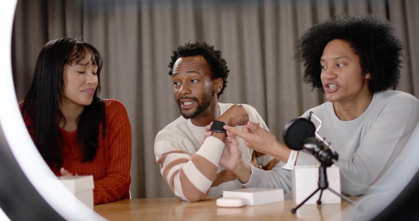 Diverse co-hosts showing smartwatch at studio table, phone showing livestream sparking demo