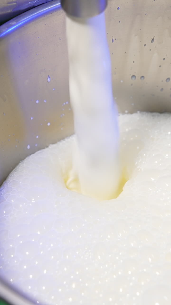 Fresh milk is pouring into a metal bucket creating foam on top. Close up. Food factory production concept. Vertical video