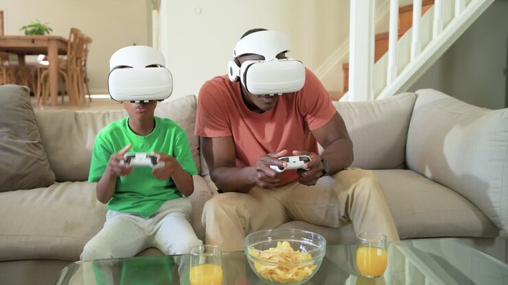 African American dad and son putting on white VR headsets, holding controllers on couch, playing VR
