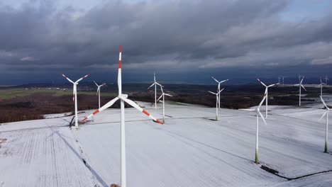 aerial high angle circling shot of spinning wind turbines working on snow-covered rural fields during sunny day with clouds in winter