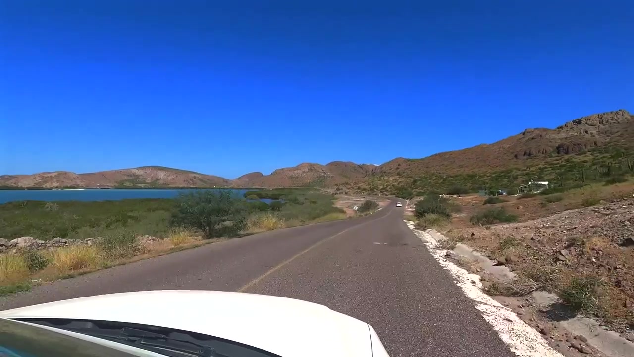 バハ・カリフォルニア・スル州の砂漠の道を小さな丘を上る車からの車窓越しの眺め。プラヤ・バランドラに近づくにつれて、海岸線沿いの左手に穏やかな入り江が見える。