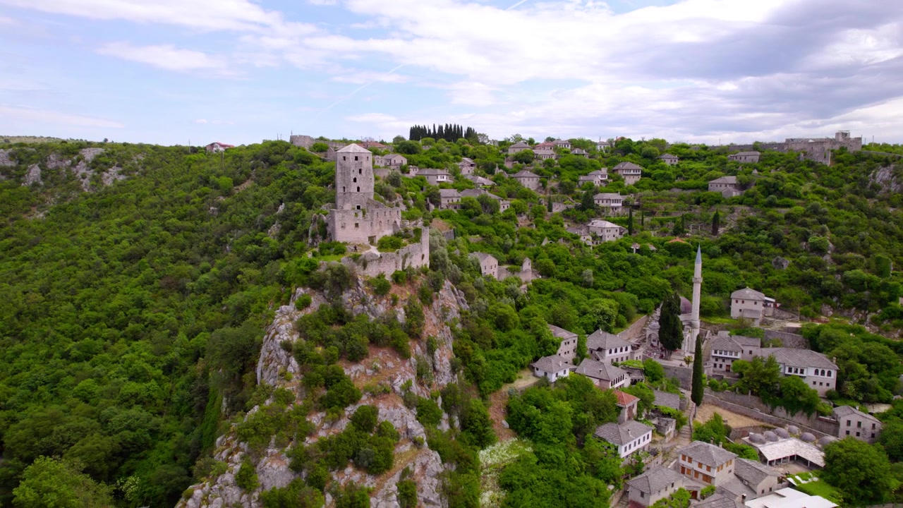 El histórico pueblo de Počitelj y su torre de piedra se elevan sobre colinas verdes en Bosnia y Herzegovina, toma aérea de establecimiento con retroceso.