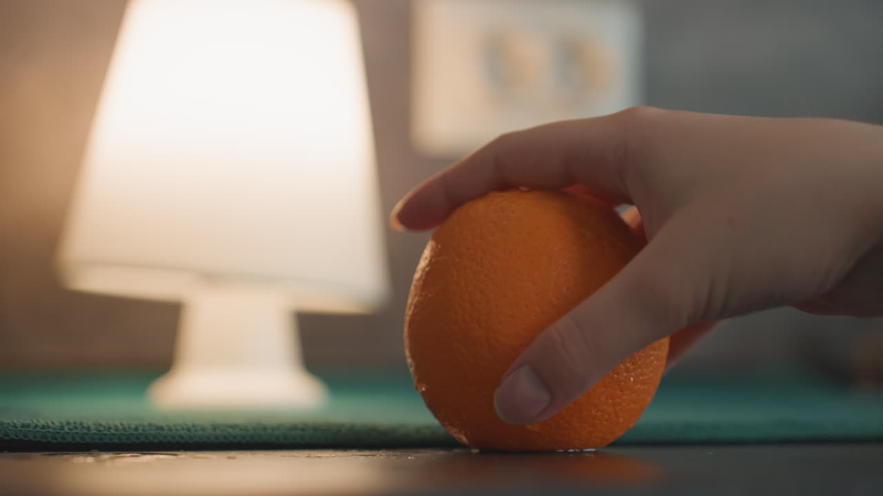 Laranja na bancada perto de uma luminária, mão se aproxima e depois espreme, gota de suco transparente cai, movimento sutil capturado, luz ambiente quente, profundidade rasa, detalhe tátil da casca, lanche experimental tranquilo.