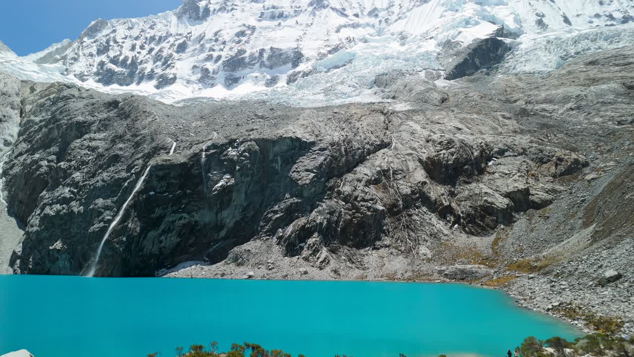 A stunning aerial shot tilts up from the vibrant turquoise water of Laguna 69 to reveal the majestic, snow-covered Chacraraju mountain peak in the Andes of Peru