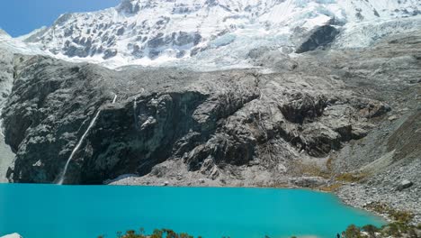 a stunning aerial shot tilts up from the vibrant turquoise water of laguna 69 to reveal the majestic, snow-covered chacraraju mountain peak in the andes of peru