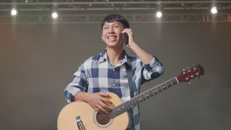 adolescente asiático con guitarra hablando en teléfono inteligente mientras está de pie en el escenario