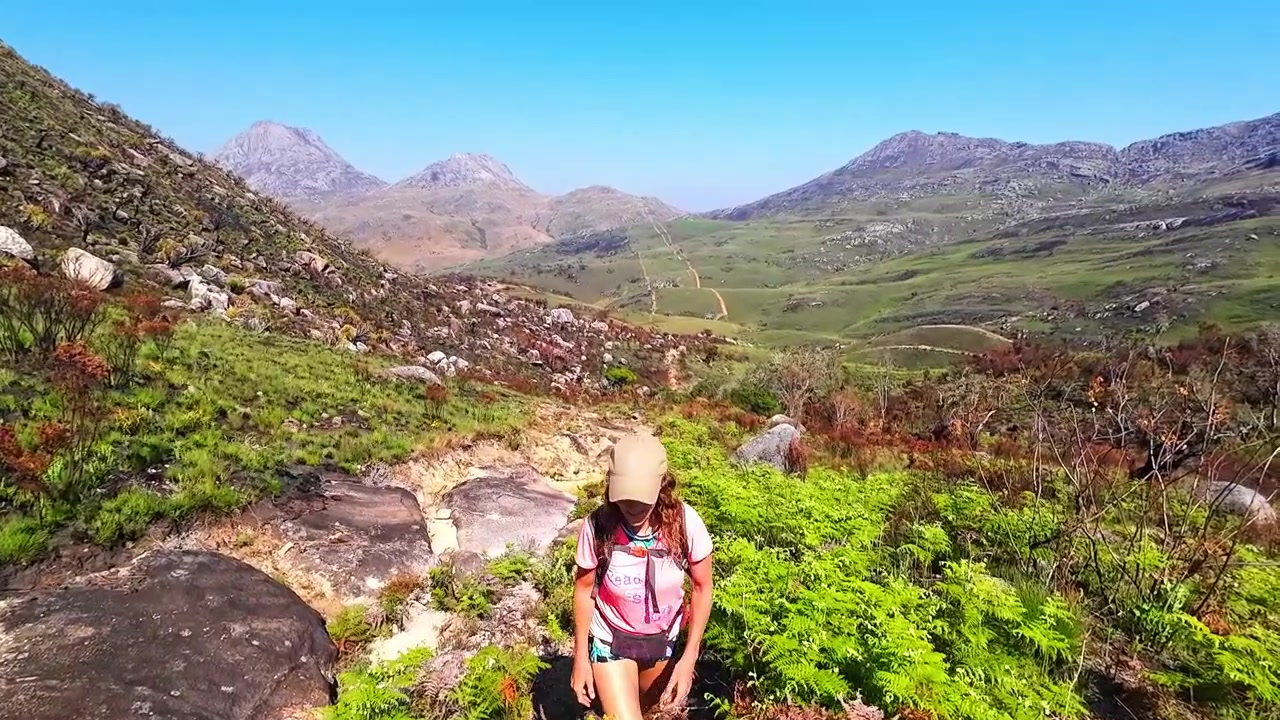 Una mujer con mochila sube por un sendero rocoso en el monte Mulanje, Malaui, rodeada de vegetación verde y escarpados picos montañosos