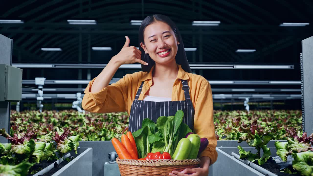 채소 바구니를 가진 아시아 여성 농부 따뜻하게 미소 짓고 스마트 농장에서 전화 제스처를 하고 있습니다.