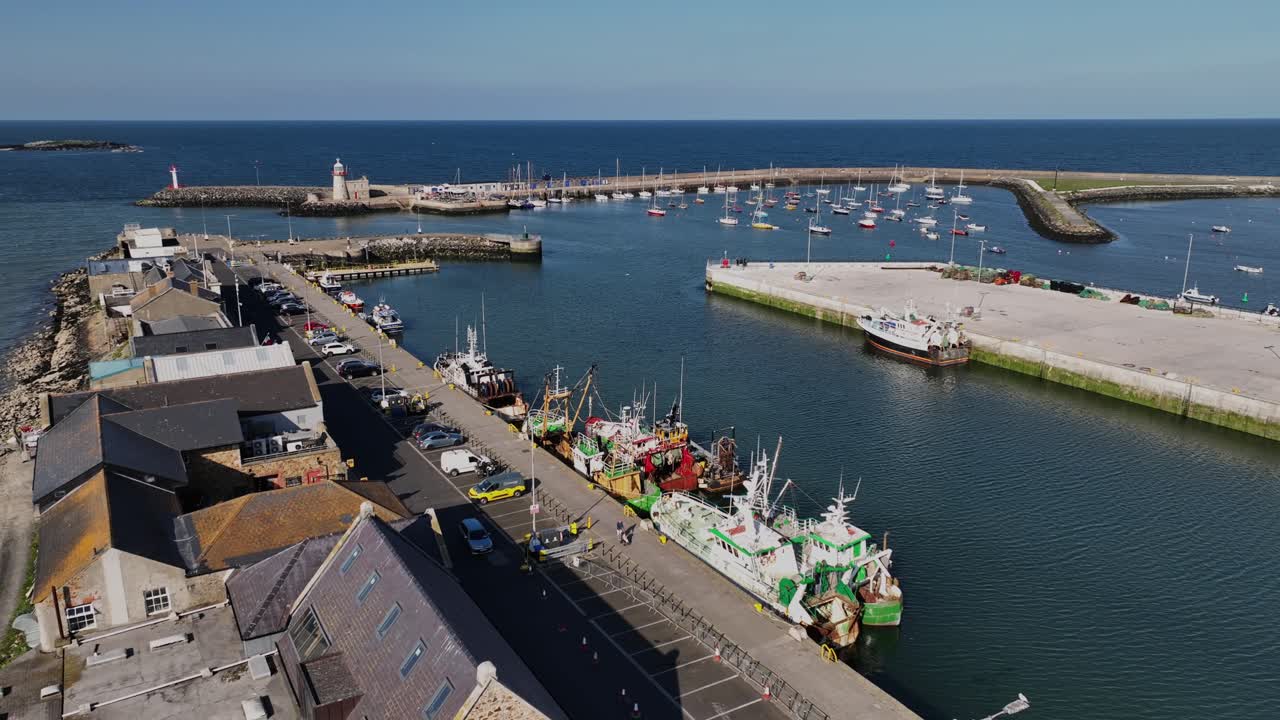 Howth Harbour, County Dublin, Ireland, May 2023. Drone slowly pulls backwards along the quayside above moored Fishing trawlers with Howth Pier and Lighthouse in the background on a warm sunny day.