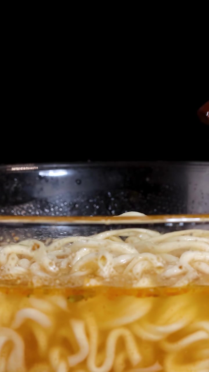 Bacchette sollevano noodles istantanei cotti da una ciotola di vetro fumante di brodo. Primo piano con illuminazione drammatica e profondità di campo ridotta, che enfatizza la consistenza e il calore.