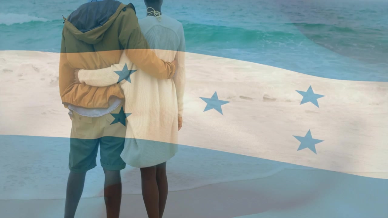 Pareja caminando por la orilla, deteniéndose, abrazándose y observando el océano bajo la bandera de Honduras ondeando, para un anuncio de viajes.