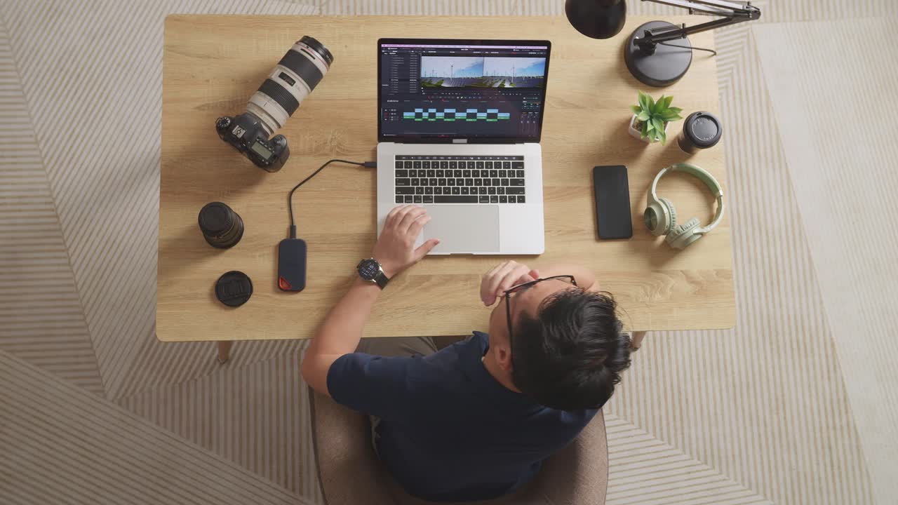 vista superior del editor masculino asiático bostezando y durmiendo mientras está sentado en el espacio de trabajo usando una computadora portátil junto a la cámara editando el video en casa