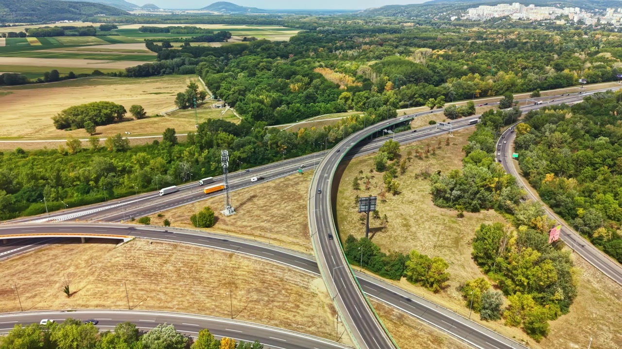 Il traffico scorre lungo un'autostrada tortuosa con campi verdi e foresta sullo sfondo vicino a una città durante una giornata di sole