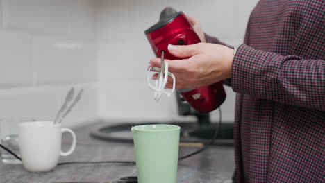 Woman using electric hand mixer inside green cup, pulling whisk out of cup after mixing, placing it in sink for cleaning, kitchen countertop with utensils and sink in background