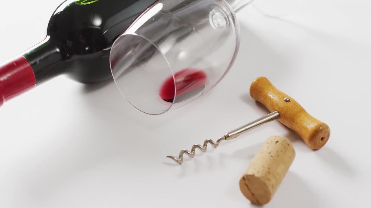 Bottiglia di vino rosso, bicchiere vuoto, tappo di sughero e cavatappi appoggiati su superficie bianca con spazio per il testo