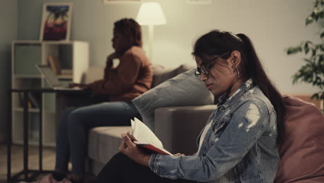 Indian-woman-reads-book-while-girl-friend-studying-on-laptop