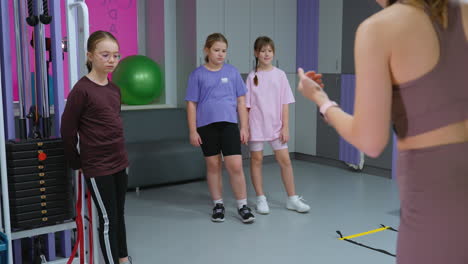 vista trasera de un instructor dirigiendo a una joven mientras otros miran atentamente, con uno descansando en un levantador de pesas, la sesión tiene lugar en un gimnasio equipado con varias herramientas de fitness