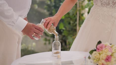 Bride-and-groom-pouring-sand-from-two-separate-vessels-into-one-vase-during-the-wedding-ceremony-on-warm-sunny-day,-close-up-shot-of-a-Unity-Sand-Ceremony