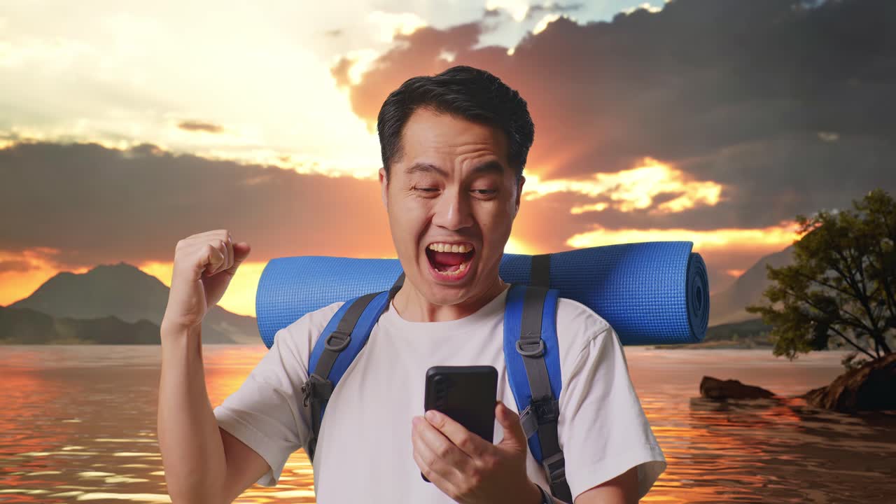 Close Up Of Asian Male Hiker With Mountaineering Backpack Looking At The Smartphone And Screaming Goal Celebrating The Success At A Lake