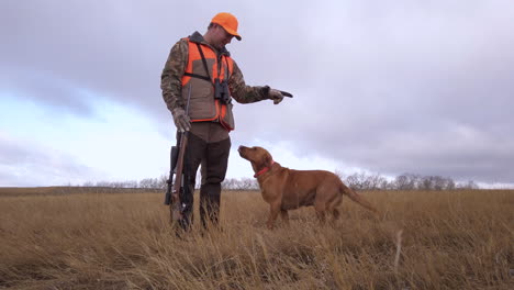 Young-Caucasian-male-hunter-standing-in-grasslands-with-hunting-dog,-petting-and-instructing-to-sit-on-cloudy-day,-Saskatchewan,-Canada,-low-angle-static-close-up