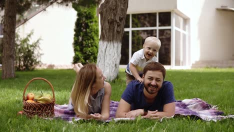 Happy-joyful-young-family-father,-mother-and-little-son-having-fun-outdoors,-playing-together-in-summer-park,-countryside.-Mom,-Dad-and-son-on-fathers-back-junping-and-laughing.-White-house-on-the-background.-Slow-motion