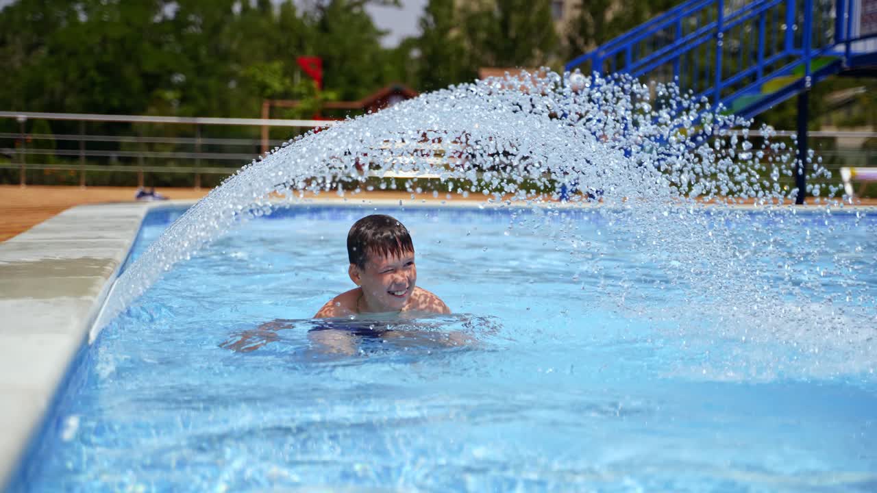 Active boy enjoys in the swimming pool. Happy child having fun near small waterfall inside the pool outdoors. Happy summertime.