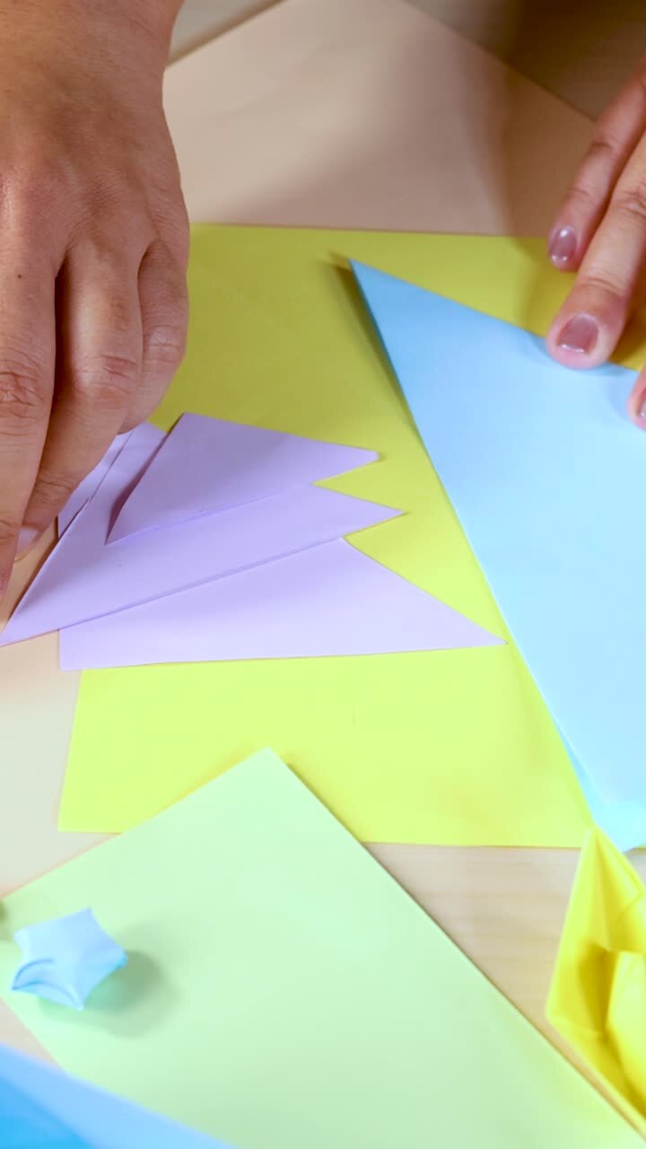 Una persona piega carta pastello in forme di origami su una scrivania, con luce intensa e angolazione della telecamera dall'alto.