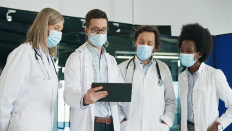 Multiethnic-doctors-team-in-medical-mask-discussing-coronavirus-disease-while-typing-on-tablet-standing-in-hospital