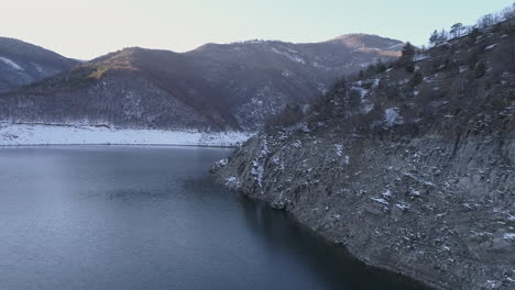 AERIAL:-Flying-above-high-rocky-shore-of-a-mountain-lake-on-its-shores-there-are-hills-covered-with-snow,-which-are-reflected-in-the-waters-of-the-lake