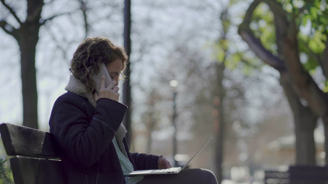 Cheerful mature woman sitting on bench and talking on smartphone