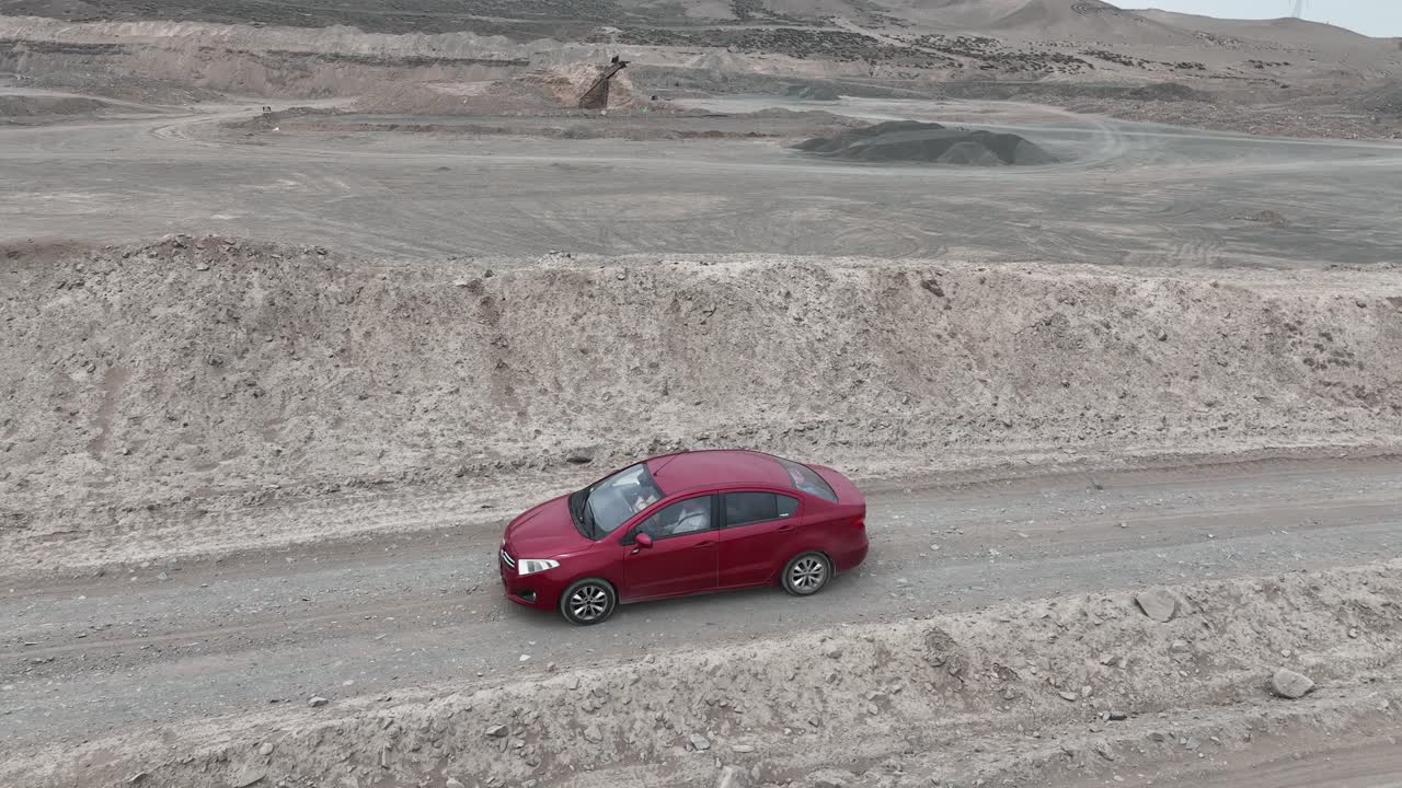 Aerial panning shot of a red vehicle driving along a dusty and stony road in the desert along stone hills in front of the holy city of Caral