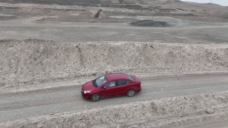 Toma-Panorámica-Aérea-De-Un-Vehículo-Rojo-Conduciendo-Por-Un-Camino-Polvoriento-Y-Pedregoso-En-El-Desierto-A-Lo-Largo-De-Colinas-De-Piedra-Frente-A-La-Ciudad-Santa-De-Caral