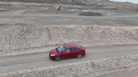 toma panorámica aérea de un vehículo rojo conduciendo por un camino polvoriento y pedregoso en el desierto a lo largo de colinas de piedra frente a la ciudad santa de caral