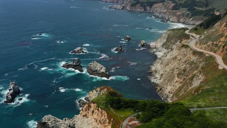 Amplia-Vista-Aérea-De-La-Sinuosa-Carretera-Con-Curvas-A-Lo-Largo-De-La-Ruta-1-En-La-Costa-De-Big-Sur-California-Con-Olas-Rompiendo-En-Las-Grandes-Rocas-En-El-Océano-Pacífico