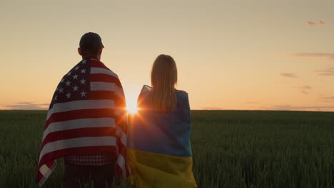 happy couple with the flags of ukraine and the usa stand side by side and look at the sunrise over a field of wheat