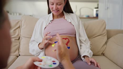 A-happy-pregnant-brunette-woman-draws-hearts-on-her-belly-with-her-husband-using-bright-colors-while-expecting-her-baby