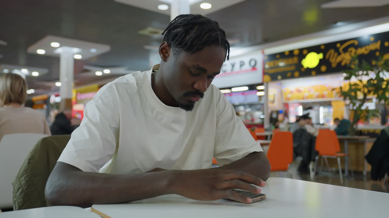 Young man in white shirt sits alone at table in indoor eatery, appearing thoughtful and reflective, surrounded by blurred background with people dining, modern decor, and illuminated signage