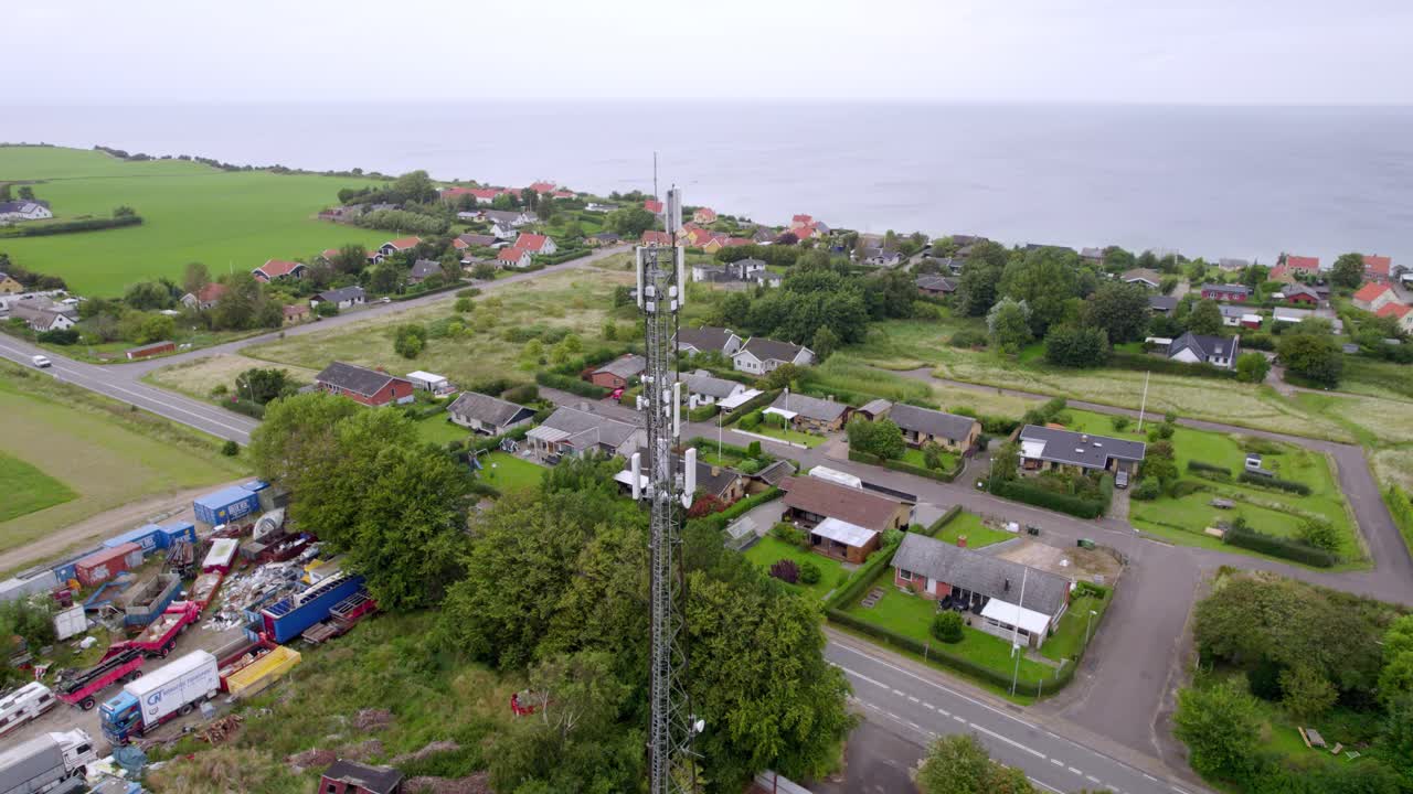 A drone captures a high angle view of a tall communication mast surrounded by houses and greenery near the Danish coastline