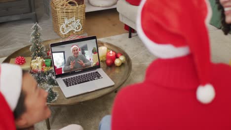 Vielfältige-Familie-Mit-Weihnachtsmützen-Nutzt-Laptop-Für-Weihnachtsvideoanruf-Mit-Glücklichem-Mann-Auf-Dem-Bildschirm